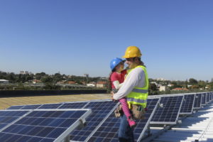 A man wearing a hardhat and safety vest holds his daughter, also in a hardhat. They are standing in a field of solar panels.