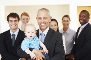 A man in a suit holds a baby in his arms and smiles at the camera while several colleagues stand behind him.