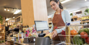 A woman is working as a checker at a grocery store. She wears an apron and is scanning items.