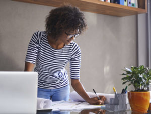 A woman leans over her desk and makes notes on an unseen paper. She is casually dressed.