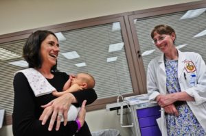 A woman holds her baby in the doctor's office while smiling at her doctor, who is nearby.