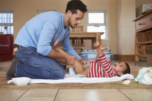 A man is at home changing his son's diaper on a changing mat on the floor.