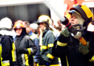 A female firefighter aims a hose at an unseen wall. The hose is spewing water and she is presumably fighting the fire. Others look on in the background.