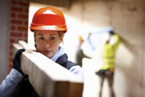 A female construction worker in a hardhat carries a large plank of wood while others work in the background.