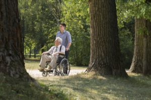 A younger man pushes an older gentleman in a wheelchair. They are walking down a path in a sunny park.