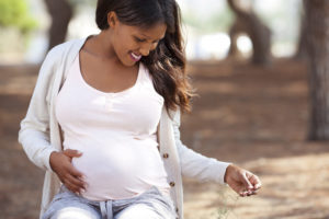 A pregnant woman smiles while sitting on a bench in the woods, holding her stomach.