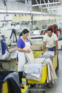 A woman folds laundry at a large laundromat. A man next to her pushes the cart of folded laundry.
