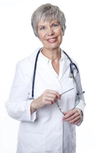 An elderly female doctor smiles at the camera. She is wearing her lab coat and a stethoscope.