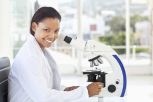 A female scientist smiles at the camera while she operates a microscope.