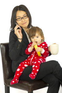 A woman in a suit holds a child in bright pajamas on her lap. She is trying to talk on her cell phone and drink a cup of coffee while holding her child.