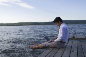 A man in a shirt and khakis sits on a dock with his feet in the water. He is on his laptop.