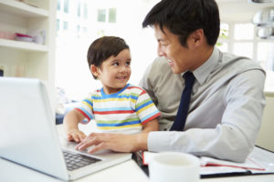 A man in a shirt and tie holds his son in his lap while working from home on his laptop. Both are smiling.