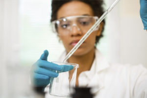 A female scientist wearing goggles and latex gloves uses a pipette to put liquid into a beaker.