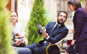 A business man in a chair outside asks a business woman behind him a question about a paper he is holding.