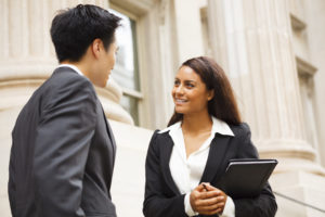 A male and female professional stand outside a columned building talking. The woman is holding a folder.