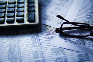 A stock photo of a keyboard, a lot of spreadsheets under it, and a pair of glasses on top of the spreadsheets in the corner.
