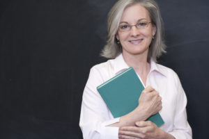 An older woman clutches a book to her chest and smiles at the camera. She is wearing a collared shirt.
