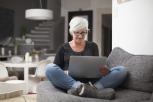 An older woman sits cross-legged in an armchair while looking at her laptop.