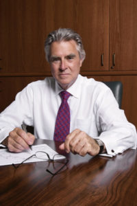 An older man wearing a shirt and tie sits at his desk and looks sternly at the camera.
