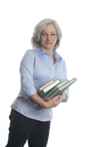An older woman holds a stack of books and looks at the camera.