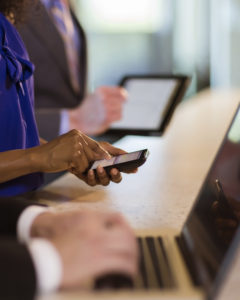 Two businessp eople use their electronic devices. One is on a phone and the other on a laptop. Only their hands and bodies are visible.