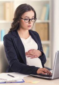 A young pregnant woman in glasses is using her laptop while working in an office.