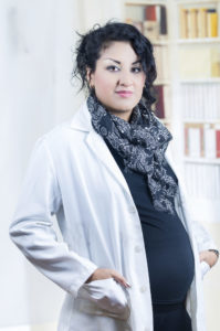 A pregnant woman is wearing a lab coat and smiling at the camera.