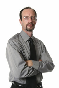 A man in a shirt and tie crosses his arms and looks sternly at the camera.