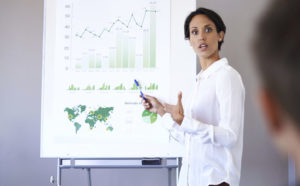 A businesswoman is explaining data to her colleagues during a meeting. She is standing in front of an easel which displays the data.
