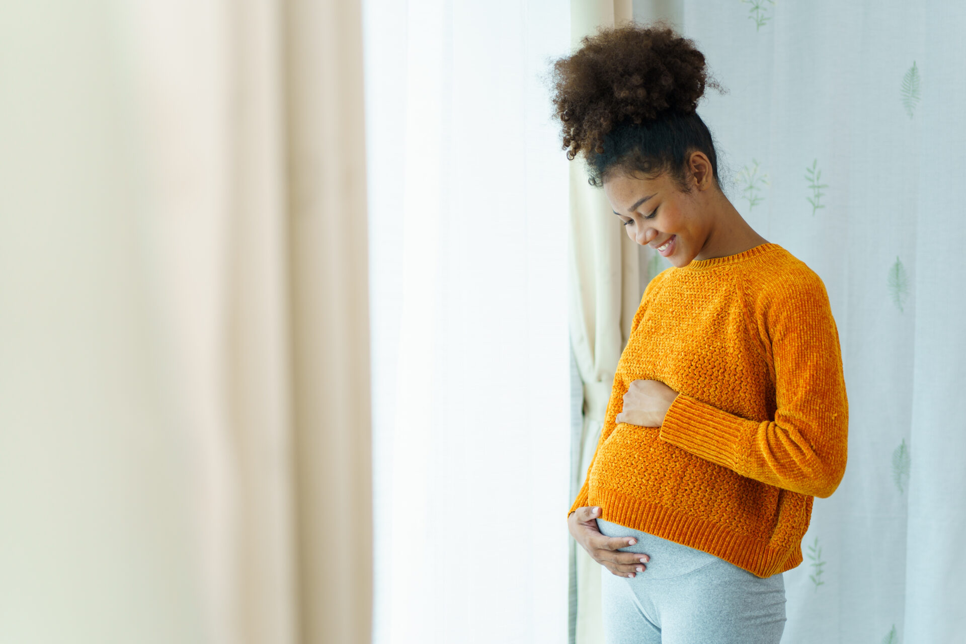 Happy cheerful African - American black pregnant woman standing near the window in bedroom and enjoy looking a natural view outside close up with copyspace. Pregnant woman portrait.